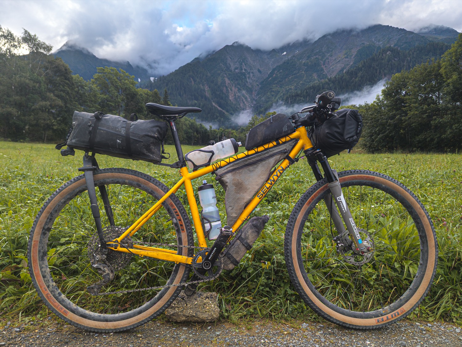 A loaded bikepacking bike in a green alpine meadow below misty peaks.